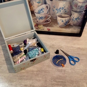 Può includere: Una scatola di legno blu chiaro piena di accessori per cucire, tra cui bobine di filo, bottoni, aghi e un metro a nastro. La scatola è decorata con un motivo floreale. C'è anche un piccolo contenitore di aghi e un paio di forbici blu su una superficie grigia.