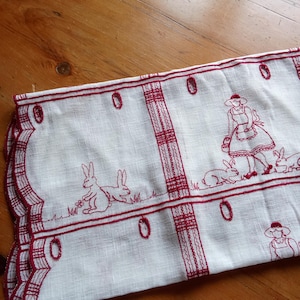 May include: White linen table runner with red embroidered design. Features a repeating pattern of rabbits, a woman in a bonnet, and decorative oval shapes. The edges are scalloped with red stitching.