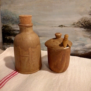 May include: Two brown ceramic containers with cork stoppers. The container on the left is taller and narrower than the container on the right. The container on the right has a small wooden spoon inside.
