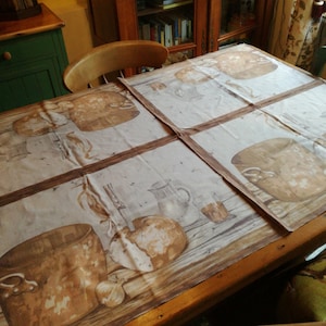 May include: A brown wooden table with a white tablecloth featuring a repeated pattern of brown pots and other kitchen items. The tablecloth is folded in the middle, creating a grid pattern.