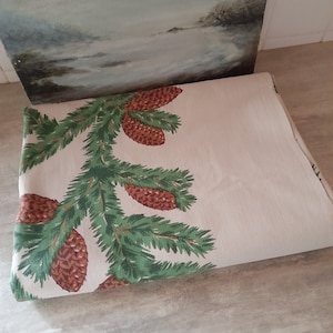 May include: A beige fabric with a repeating pattern of green pine branches and brown pine cones. The fabric is folded in half and lying on a wooden surface.