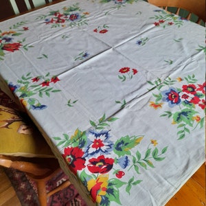 May include: A vintage floral tablecloth with a light blue background and a white border. The tablecloth features a repeating pattern of red, yellow, blue, and green flowers.