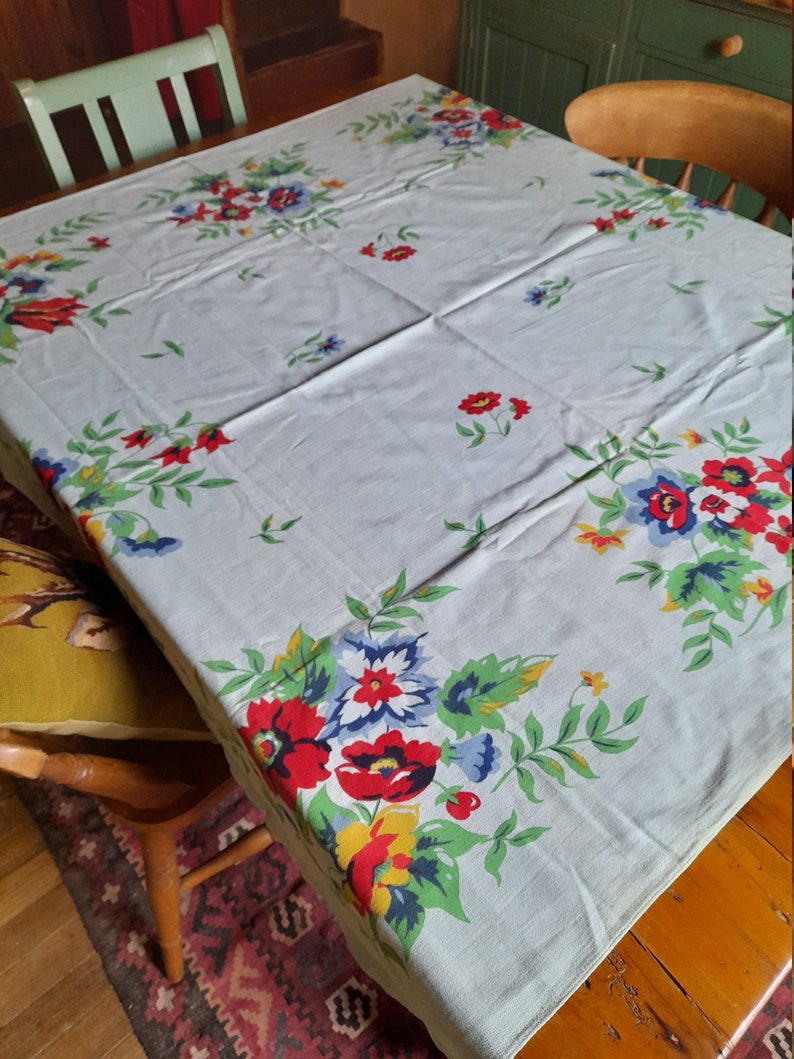 May include: A vintage floral tablecloth with a light blue background and a white border. The tablecloth features a repeating pattern of red, yellow, blue, and green flowers.