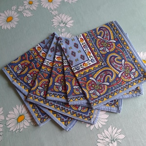 May include: A stack of six blue paisley patterned cloth napkins. Each napkin features a detailed design with red, yellow, and white floral elements. The napkins are arranged on a light green surface with a daisy pattern.