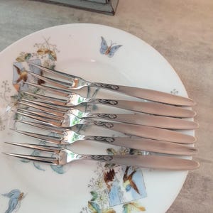 May include: A set of six silver forks arranged on a white plate with a floral and butterfly design. The forks have a decorative pattern on the handles. The plate has a delicate, vintage aesthetic, suggesting a formal dining setting.