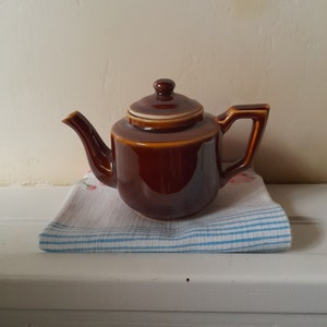 Puede incluir: Una tetera de cerámica marrón con tapa, sentada sobre una toalla de té a rayas blancas y azules.