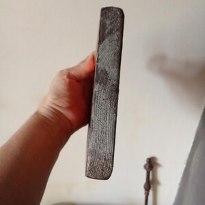 May include: A rectangular block of dark, weathered wood with a rough, textured side surface showing visible grain patterns. The object has a solid, dense appearance with a worn, matte finish and straight edges, presented in a vertical orientation.