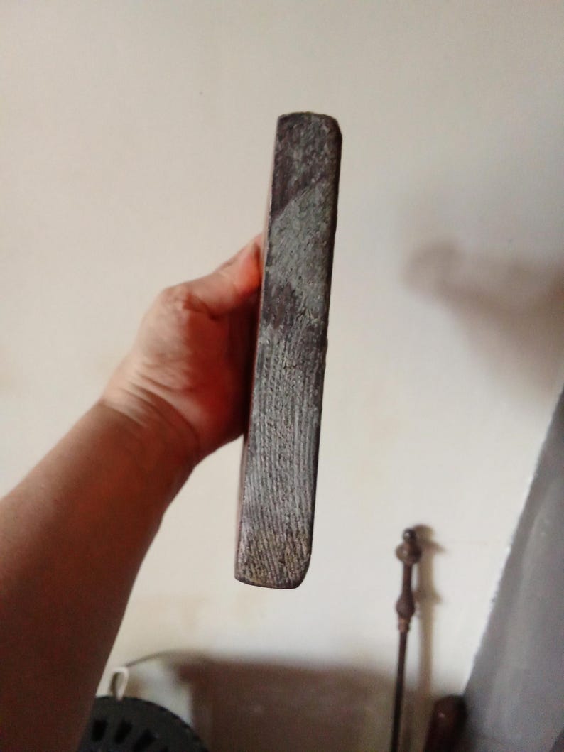 May include: A rectangular block of dark, weathered wood with a rough, textured side surface showing visible grain patterns. The object has a solid, dense appearance with a worn, matte finish and straight edges, presented in a vertical orientation.