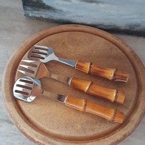 Puede incluir: Tres utensilios de servicio de plata con mangos de bambú sobre una tabla de cortar de madera. Los utensilios tienen una cuchara ranurada, un tenedor y una espátula.