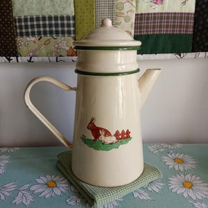 以下が含まれることがあります： クリーム色のエナメルコーヒーポット、緑色のトリムと蓋付き。 ポットにはハンドルと注ぎ口があり、小さなフェンスの近くに2匹のウサギのグラフィックがあります。 緑色の正方形の鍋敷きの上に置かれています。