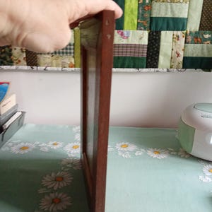May include: A rectangular, dark brown wooden picture frame. The frame is held by a hand, showcasing its side profile. The frame is empty, ready for a photo or artwork. The background features a floral tablecloth and a patchwork quilt.