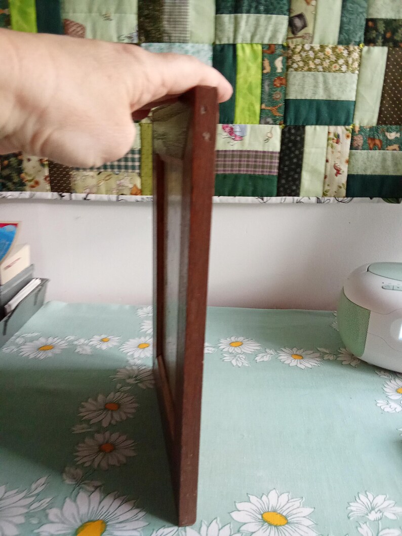 May include: A rectangular, dark brown wooden picture frame. The frame is held by a hand, showcasing its side profile. The frame is empty, ready for a photo or artwork. The background features a floral tablecloth and a patchwork quilt.