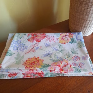 May include: A stack of folded white fabric with a floral pattern in shades of pink, red, yellow, and blue. The fabric has a white background with a variety of flowers and leaves.