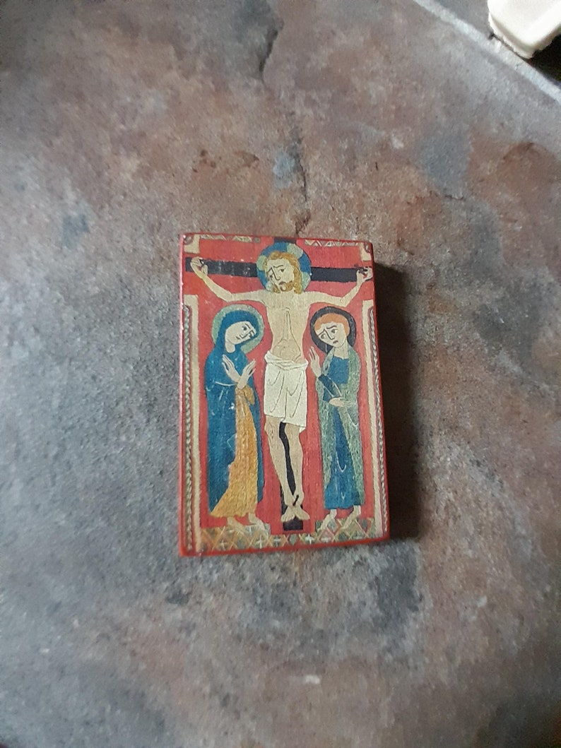 May include: A small, rectangular wooden panel with a painted depiction of the crucifixion of Jesus Christ. The panel is painted in a red, blue, and yellow color scheme. The figures are stylized and have a medieval appearance.