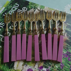 Peut inclure: Ensemble de fourchettes anciennes avec des dents dorées et des manches roses. Les fourchettes sont disposées en rangée, mettant en valeur leur design élégant. Les manches sont d'un rose uni et doux, contrastant avec les dents dorées.