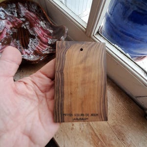 May include: A small, rectangular wooden plaque with the text "Petites Soeurs de Jesus Jerusalem" engraved on the bottom. The plaque is made of dark wood with a natural grain pattern.