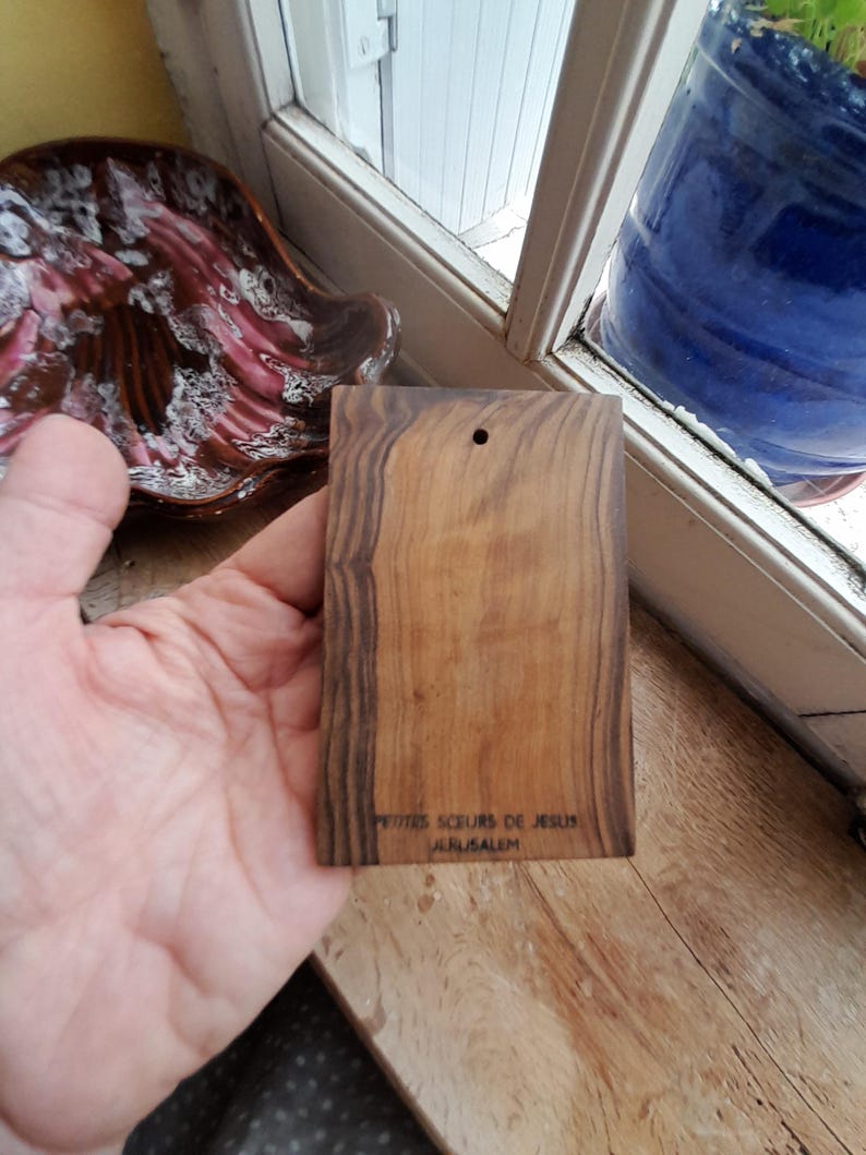 May include: A small, rectangular wooden plaque with the text "Petites Soeurs de Jesus Jerusalem" engraved on the bottom. The plaque is made of dark wood with a natural grain pattern.