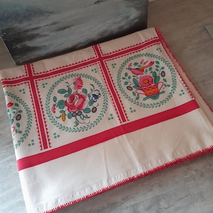 Könnte beinhalten: Weiße Tischdecke mit rotem Rand und einem sich wiederholenden Muster aus Blumendesigns in den Farben Rosa, Grün, Blau und Gelb. Das Design zeigt Vögel, Blumen und Blätter.
