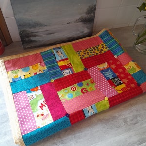 Puede incluir: Una colcha de retazos colorida con una variedad de cuadrados de tela con temática de gatos. La colcha está hecha con una mezcla de telas rosas, rojas, azules, verdes y amarillas. La colcha está doblada por la mitad y tiene un borde amarillo.