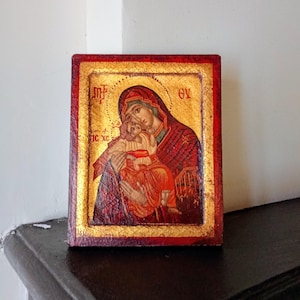 May include: A rectangular religious icon featuring a painted depiction of a figure holding a child against a gold leaf background. The border is painted in a deep red hue. Greek lettering is visible in the upper corners and near the center.