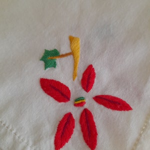 Op de afbeelding: Een witte stof met een rode geborduurde bloem met een groen blad en een gele stengel. Het midden van de bloem heeft een kleine groene, gele en rode streep.