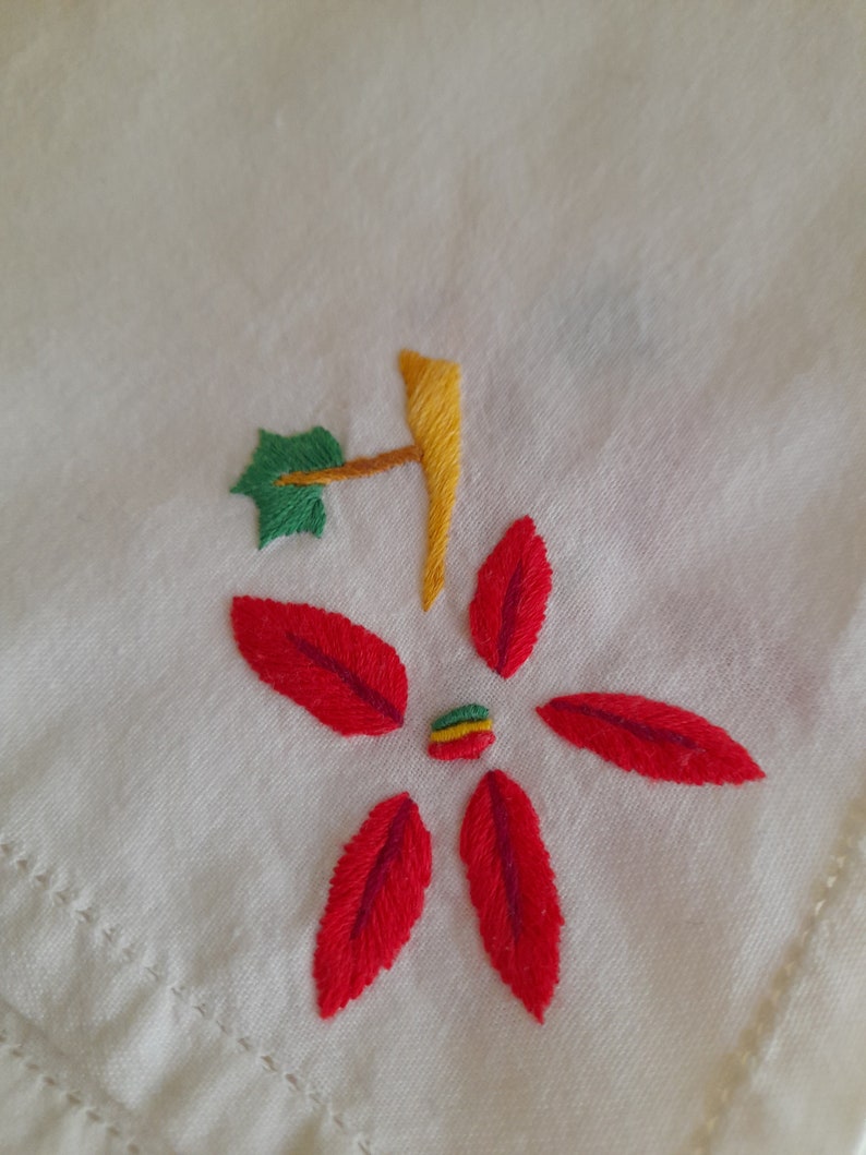 Op de afbeelding: Een witte stof met een rode geborduurde bloem met een groen blad en een gele stengel. Het midden van de bloem heeft een kleine groene, gele en rode streep.