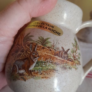 Op de afbeelding: Een beige keramische kan met een bruine en witte konijnenillustratie. De kan heeft een gespikkelde glazuur en een gouden label met de tekst "Collection Gres de la Roche".