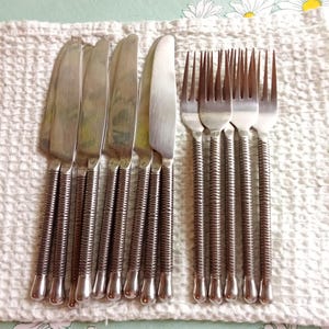May include: A collection of stainless steel knives and forks. The knives feature a rounded blade and a ridged handle. The forks have four prongs and a matching ridged handle. The cutlery is displayed on a white cloth.
