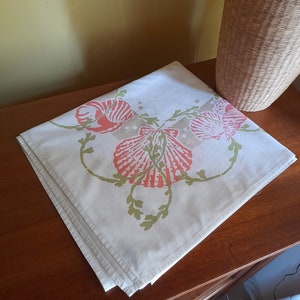 May include: A white tablecloth with a floral design featuring pink and green seashells. The tablecloth is folded and placed on a wooden surface.