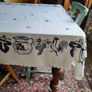Könnte beinhalten: Eine weiße Tischdecke mit einem blauen und schwarzen Muster aus Trauben, einem Hahn und einem Krug. Die Tischdecke liegt auf einem Tisch mit einem Holzbein und einem grünen Stuhl. Der Tisch steht auf einem Teppich mit einem roten und schwarzen Muster.