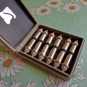 May include: A set of six silver-toned knitting needles with black bands, presented in a gray velvet-lined box. The needles have decorative finials and are arranged neatly. The box is open, revealing the contents against a floral background.