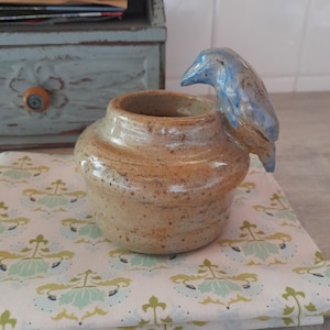 May include: A handmade ceramic jar with a blue bird perched on the rim. The jar is tan with a speckled finish, and the bird is blue. The jar sits on a white cloth with a floral pattern. A small, light blue wooden box is in the background.