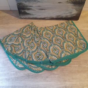 Può includere: Un set di sei tovaglioli di stoffa con motivo verde e giallo con bordo turchese. I tovaglioli sono piegati e impilati su una superficie marrone chiaro.