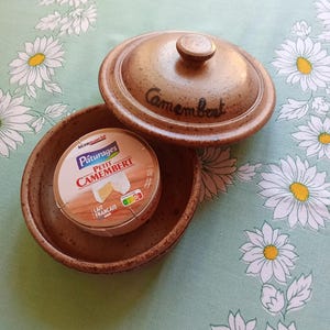 May include: A round, speckled brown ceramic cheese baker with a matching lid featuring the word Camembert written in black script. Inside the baker sits a circular wooden box of Pâturages Petit Camembert cheese with a white, red, and blue label.