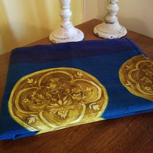 Op de afbeelding: Een gevouwen blauwe en turquoise stoffen tafelkleed met een gouden bloemenpatroon. Het tafelkleed ligt op een houten tafel met twee witte kandelaars.