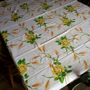K&ouml;nnte beinhalten: Wei&szlig;e Tischdecke mit einem floralen Muster aus gelben Rosen und Weizen&auml;hren. Die Tischdecke liegt auf einem Holztisch mit zwei St&uuml;hlen.