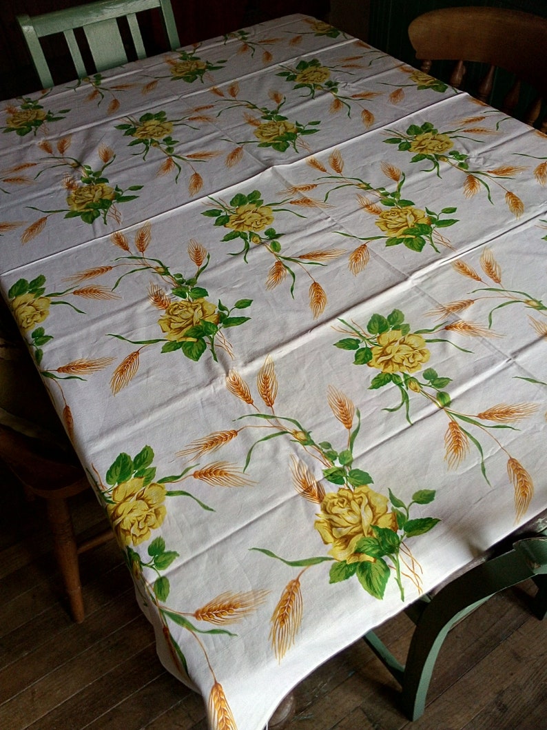 K&ouml;nnte beinhalten: Wei&szlig;e Tischdecke mit einem floralen Muster aus gelben Rosen und Weizen&auml;hren. Die Tischdecke liegt auf einem Holztisch mit zwei St&uuml;hlen.