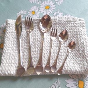 Puede incluir: Un juego de seis utensilios plateados, que incluyen un cuchillo, tenedores y cucharas, dispuestos sobre un paño blanco texturizado. Los utensilios tienen un mango en forma de lágrima y están sobre un fondo floral verde claro.