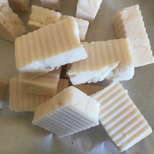May include: A collection of handmade soap bars, cut into rectangular shapes. The soap has a light beige color and a textured, ridged surface on one side. The bars are scattered, suggesting a natural, rustic aesthetic.