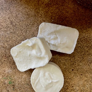 May include: Three pieces of white soap, two rectangular and one round, are arranged on a textured, brown surface. The soap has a slightly uneven texture, suggesting a handmade quality. The lighting highlights the subtle variations in the soap's surface.