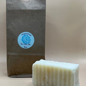 May include: A bar of tea tree oil soap next to a brown paper bag. The soap is rectangular, with a light color and visible ridges. The bag has a blue label with the text "Attagirl Soaps Tea Tree Oil Bar".