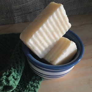 May include: Two rectangular bars of handmade soap, light cream in color, rest in a small blue and white striped bowl. The soap has a ridged texture. A green crocheted washcloth is visible in the lower left corner.