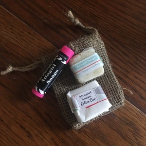 May include: A small burlap bag containing Attagirl Soaps products. The bag holds a lip balm, a bar of classic shampoo and body bar soap, and a lotion bar. The lip balm is black and white with a pink cap. The soaps are wrapped in white paper.
