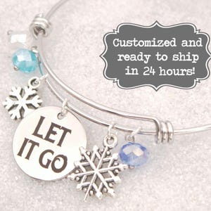 May include: A silver-toned bangle bracelet with snowflake charms and blue crystal beads. A round charm reads "LET IT GO" in black letters. The bracelet has a winter theme.