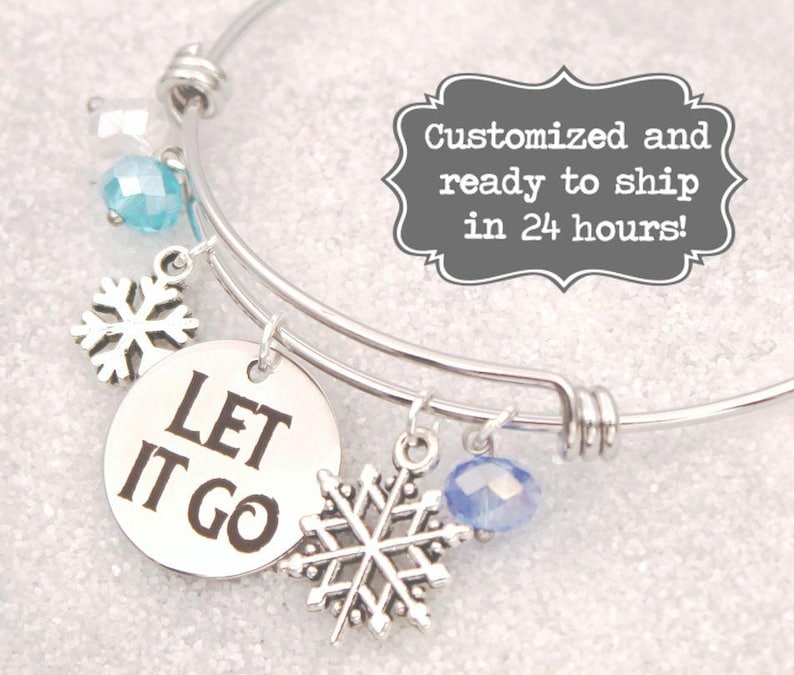 May include: A silver-toned bangle bracelet with snowflake charms and blue crystal beads. A round charm reads "LET IT GO" in black letters. The bracelet has a winter theme.