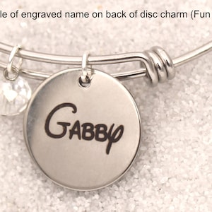 May include: A silver bangle bracelet with a silver disc charm that says "GABBY" in a fun font. The charm is attached to the bracelet with a silver ring. A small clear crystal dangles from the bracelet.