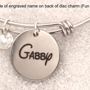 May include: A silver-toned bangle bracelet with a round charm engraved with the name "Gabby" in a playful font. A clear, faceted bead hangs nearby. The text "Example of engraved name on back of disc charm (Fun font)." is at the top.