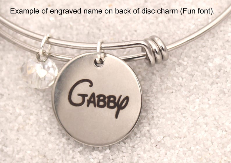 May include: A silver-toned bangle bracelet with a round charm engraved with the name "Gabby" in a playful font. A clear, faceted bead hangs nearby. The text "Example of engraved name on back of disc charm (Fun font)." is at the top.