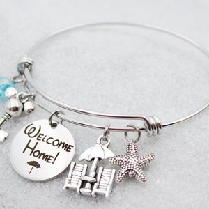 Pode incluir: Pulseira de prata com um charme redondo que diz "Bem-vindo a casa!" e outros encantos, incluindo uma chave, um cora&ccedil;&atilde;o, uma cadeira de praia, um guarda-chuva e uma estrela do mar.