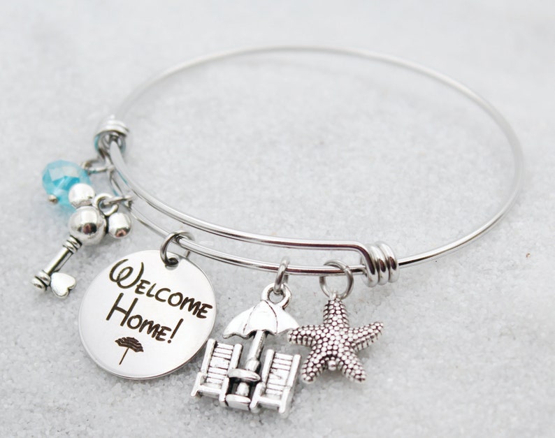 Pode incluir: Pulseira de prata com um charme redondo que diz "Bem-vindo a casa!" e outros encantos, incluindo uma chave, um cora&ccedil;&atilde;o, uma cadeira de praia, um guarda-chuva e uma estrela do mar.